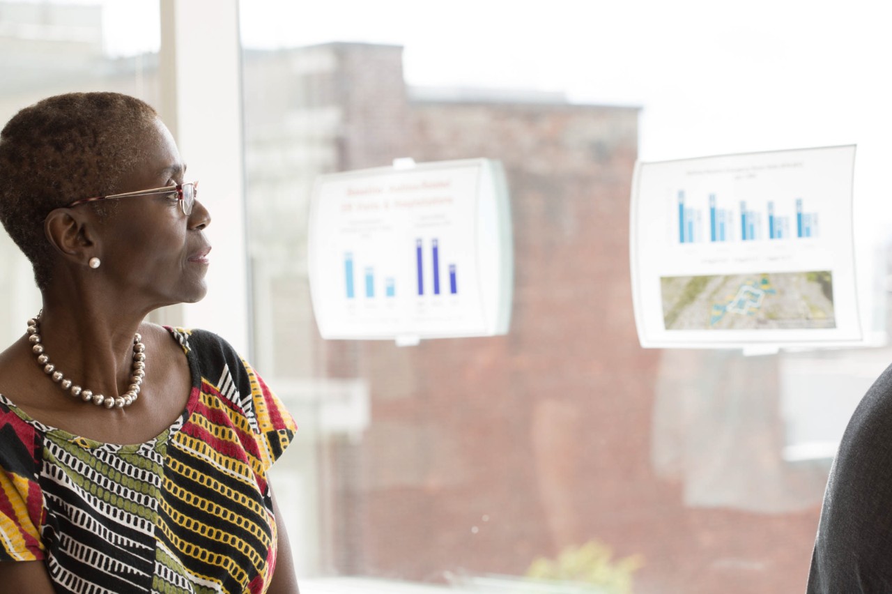 A woman refers to bar charts taped to a window while facilitating a meeting. 