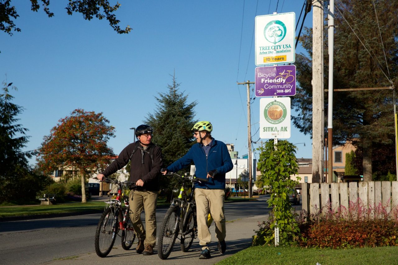 Two cyclists walk their bikes past bike-friendly signage on a downtown road. 